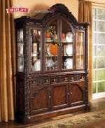 Tampilan penuh lemari hias jati mewah seri North Shore 3 pintu kaca warna cokelat gelap dihiasi pajangan cangkir kristal di dalam ruang keluarga produk Hasan Jati Furniture.