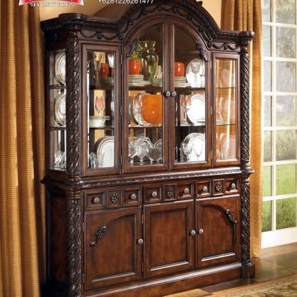 Tampilan penuh lemari hias jati mewah seri North Shore 3 pintu kaca warna cokelat gelap dihiasi pajangan cangkir kristal di dalam ruang keluarga produk Hasan Jati Furniture.