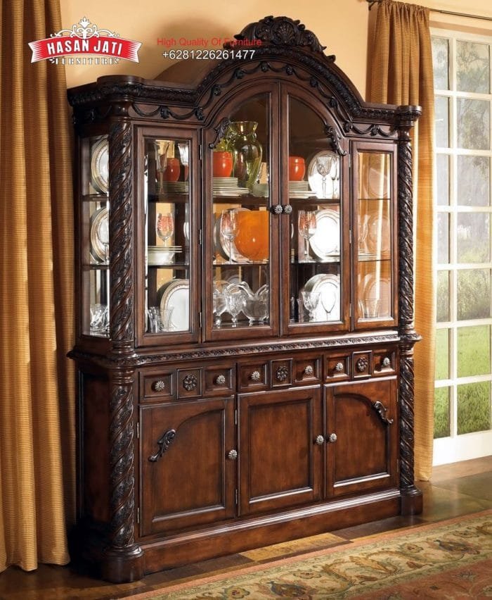 Lemari Hias Jati North Shore Mewah 3 Pintu Kaca Tampilan penuh lemari hias jati mewah seri North Shore 3 pintu kaca warna cokelat gelap dihiasi pajangan cangkir kristal di dalam ruang keluarga produk Hasan Jati Furniture.