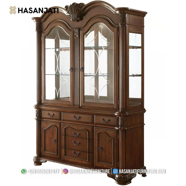 Lemari hias jati mewah tipe 2 pintu kaca seri Chateau De Ville warna cokelat berkilau dipajang di samping jendela ruang keluarga produk Hasan Jati Furniture.